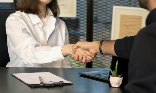 Employer and employee shaking hands across a desk over benefits paperwork