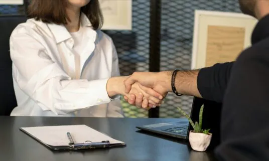 Employer and employee shaking hands across a desk over benefits paperwork